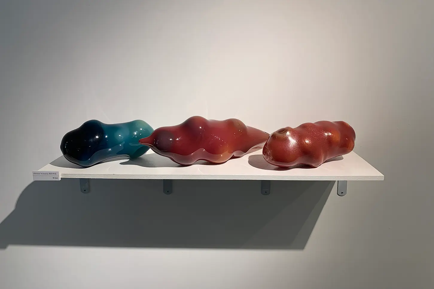 真っ白な壁に金具を介して取り付けられた白い板状の展示台がある。その上には青、赤を基調としたガラス製の作品が３つ置かれている。向かって一番左の作品は深い青、薄い青、白の3色で構成されていて、３つの作品の中で長さが一番短く小さい作品である。膨らみが4箇所ある。真ん中の作品は両橋が尖ったいて丸い膨らみが5箇所以上ある。赤と白の2色で構成されていて３つの作品の中で一番長さがある作品である。一番右の作品は両端は丸く膨らみが7箇所以上ある。真ん中の作品と色味は同じであるが三つの作品の中で一番多くの丸い膨らみがある。右上からスポットライトが照らされており影が左側に寄って落ちている。