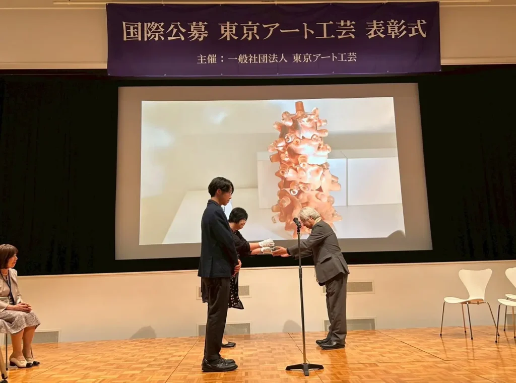 三田村審査員賞の授賞式の様子。壇上に野田紘が立っており、三田村審査員が女性からトロフィーを受け取って、野田紘に渡そうとしている。背面スクリーンに野田紘の受賞作が映し出されている。