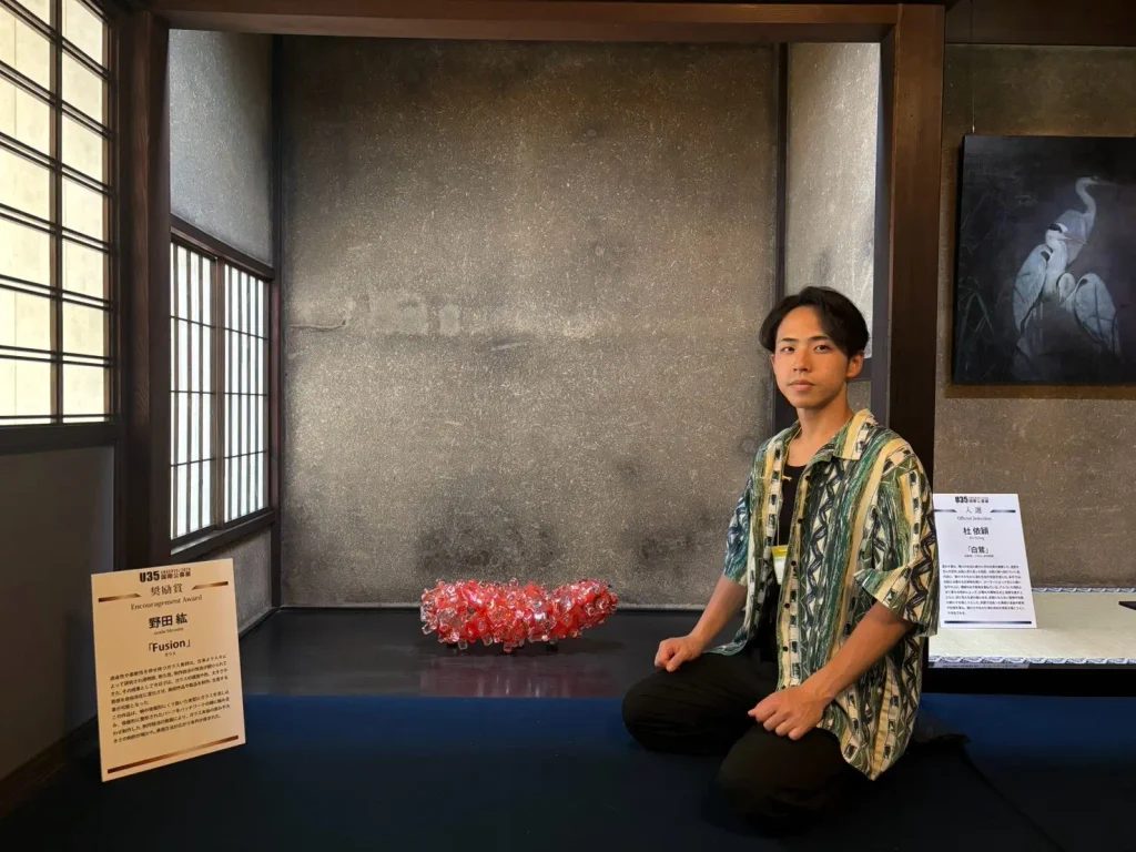 京都・大徳寺黄梅院で開催された第4回日本国際芸術祭「U35 国際公募展」にて、奨励賞を受賞した赤いガラス作品《Fusion》と、その隣に座るガラス作家・野田紘。歴史ある和室の展示空間で撮影された記念写真。